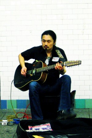 A New York City musician playing guitar in the Times Square subway station in NY. Photo Credit: Mo Riza. Photo Courtesy of: Wikimedia Commons.